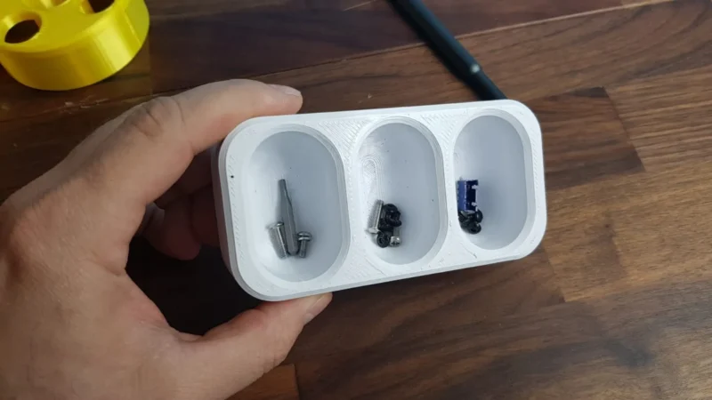 A hand is seen holding a small three compartment white organizer with screws and other small items in it. 